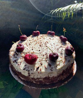 A cake finished with cream, dark chocolate shavings and whole cherries.
