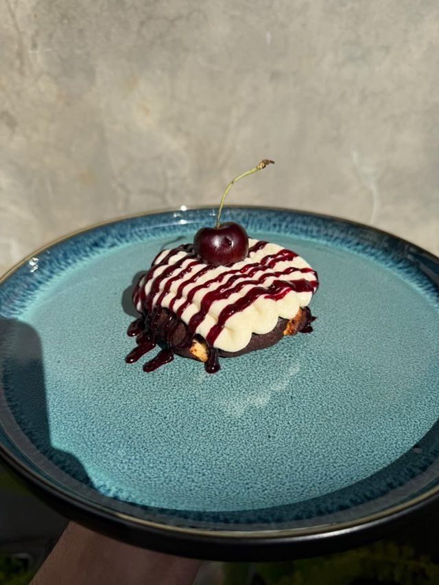A dark chocolate cookie under piped cream and cherry drizzle, topped with a cherry, on a teal plate.