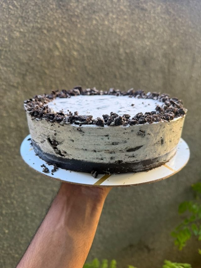 A cookies-and-cream cake edged with crushed chocolate cookies, held against a textured wall.