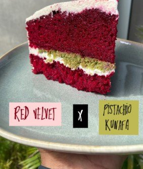 A tall slice of red velvet cake layered with pistachio kunafa, on a pale blue plate.